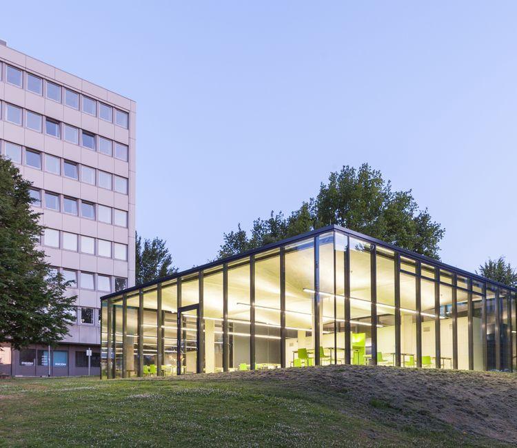 Verglaster Lernpavillon der RWTH Aachen bei Dämmerung, mit beleuchtetem Innenraum und angrenzendem Hochschulgebäude im Hintergrund.