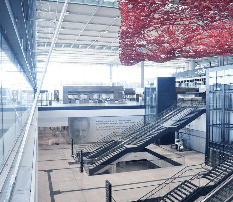 Blick durch Glaswand auf BER-Terminal mit Rolltreppen, Kunstinstallation und Zitatwand.