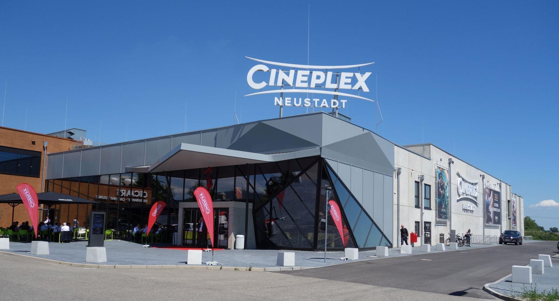 Modernes Cineplex-Kino in Neustadt mit markanter Glasfassade, großem Eingangsbereich und Außenbestuhlung. Werbeplakate und Fahnen schmücken den Vorplatz.