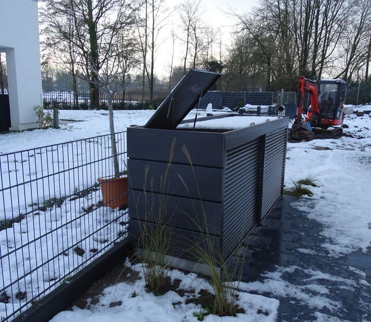 Müllbox mit geöffnetem Deckel in einem verschneiten Garten mit moderner Bebauung.