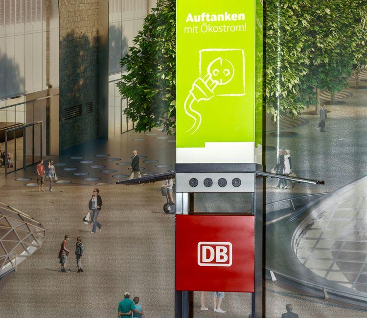 Nahaufnahme einer DB-Ladesäule mit Ökostrom-Werbung vor Wandbild eines Bahnhofs