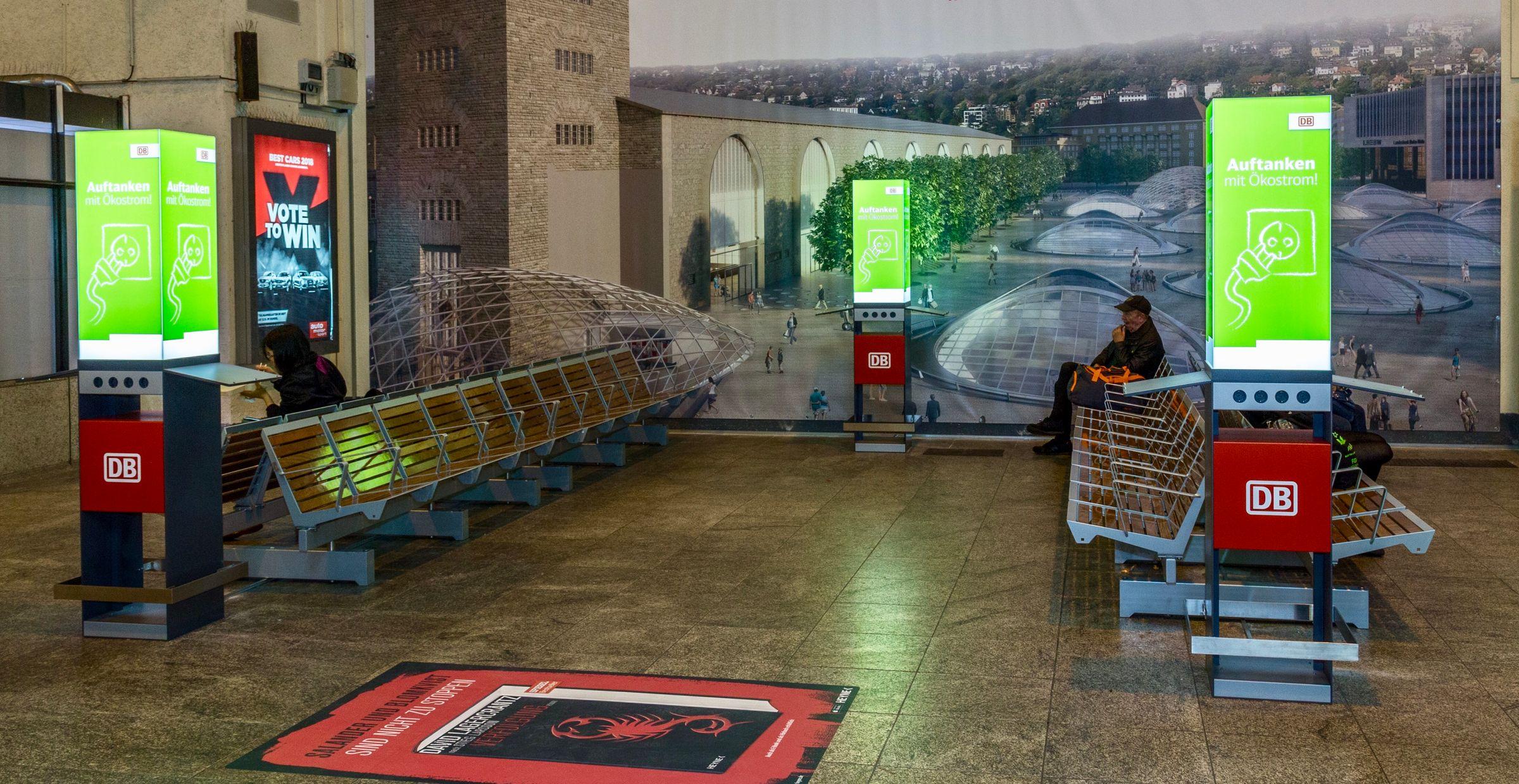 Drei DB-Ladesäulen mit Sitzbänken im Bahnhof, Werbespruch: „Auftanken mit Ökostrom!“