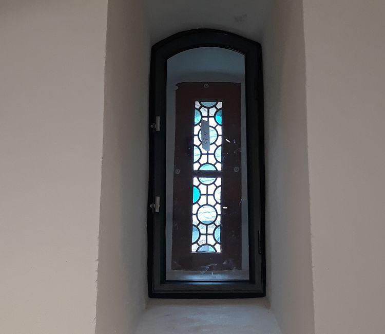 Innenansicht eines kleinen Rundbogenfensters mit historischen, farbigen Bleiglasmustern.