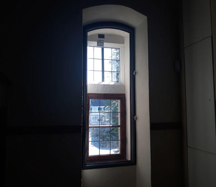 Modernisiertes, schmal hohes Fenster in einer historischen Mauer mit Blick auf ein Originalfenster aus Holz.