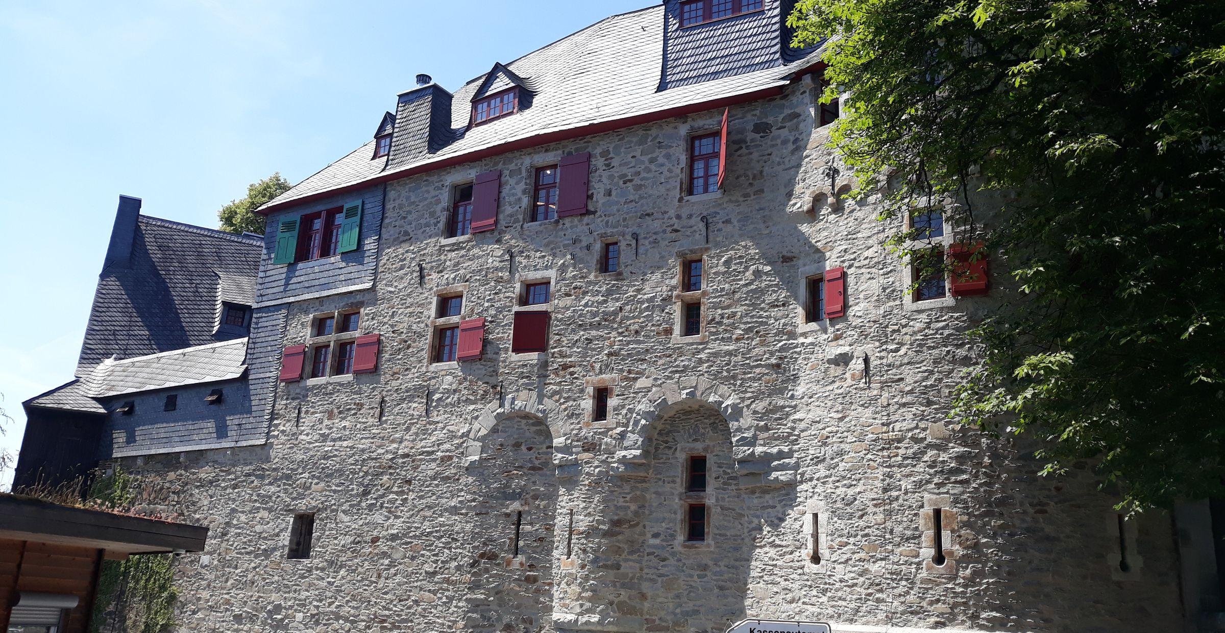 Seitenansicht von Schloss Burg mit massiver Natursteinfassade, roten Fensterläden und Fahrzeugen im Vordergrund.