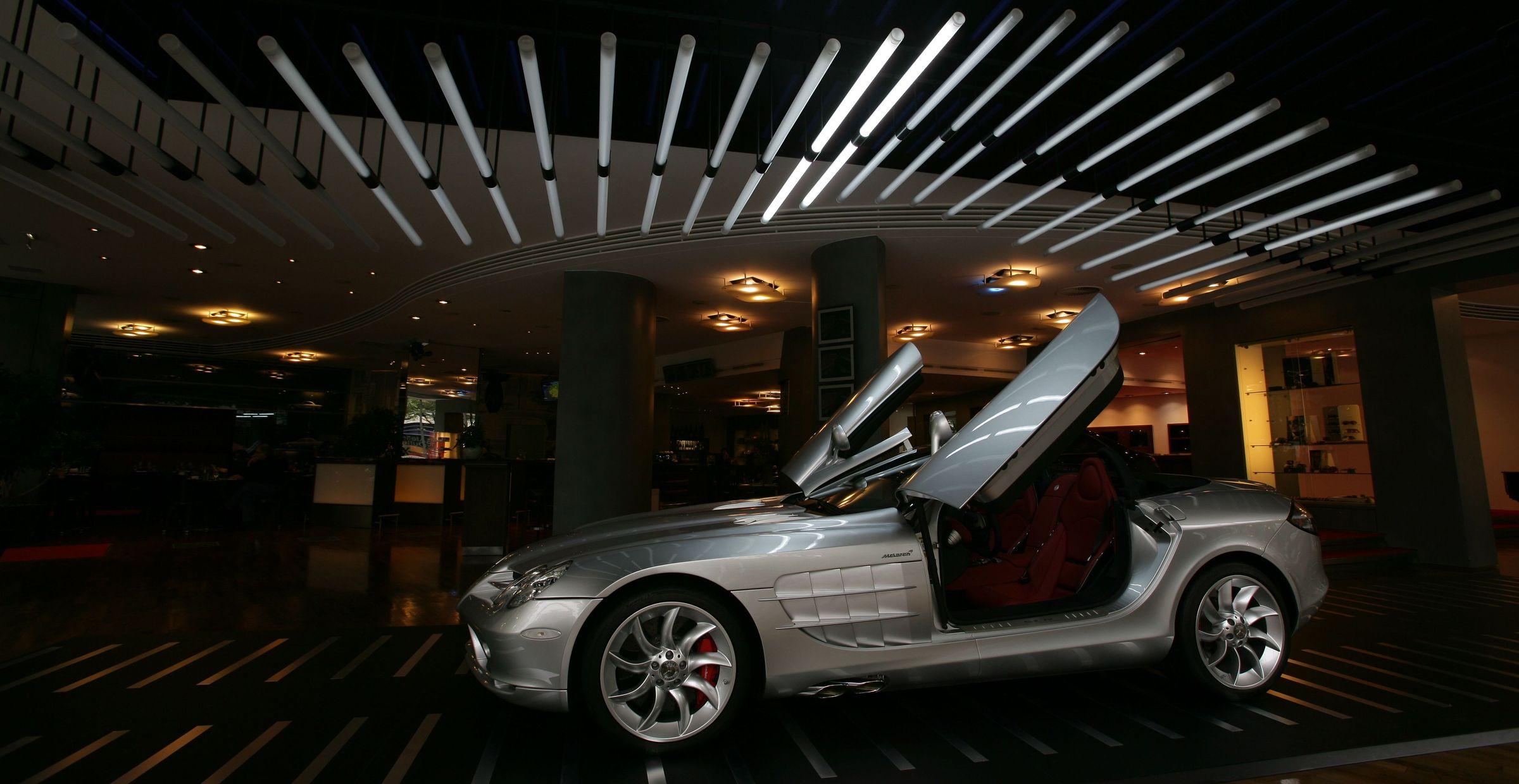 Silberner Sportwagen mit geöffneten Flügeltüren in einem modern beleuchteten Showroom mit markanter Lichtinstallation an der Decke.
