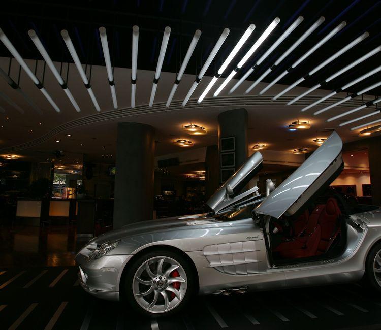 Silberner Sportwagen mit geöffneten Flügeltüren in einem modern beleuchteten Showroom mit markanter Lichtinstallation an der Decke.