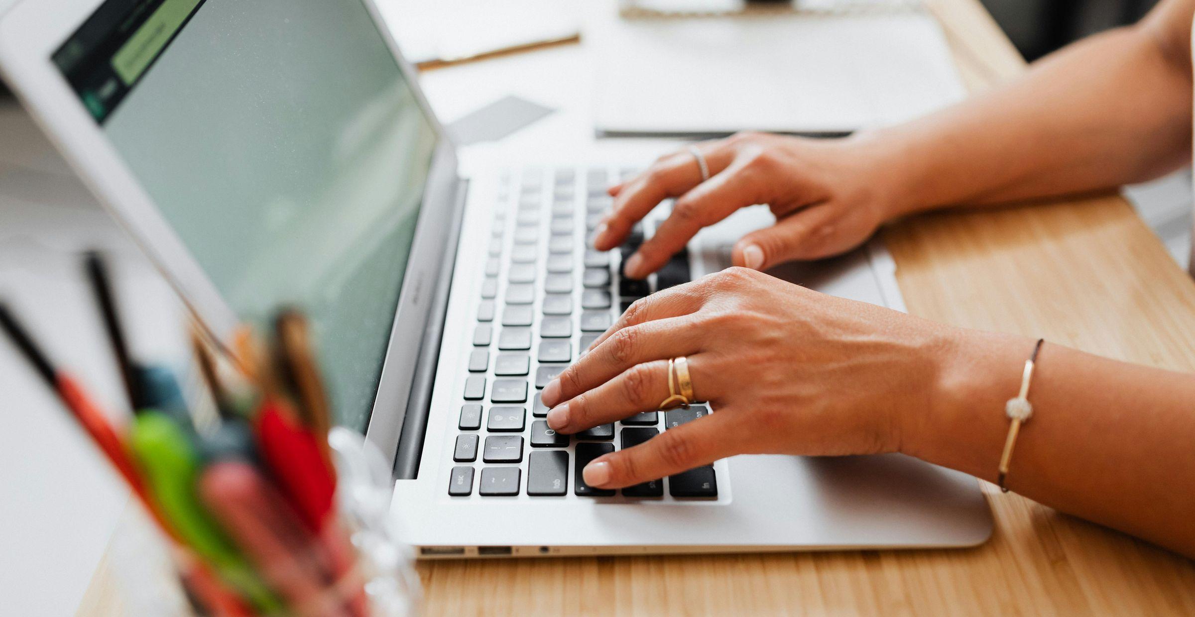 Person tippt auf Laptop-Tastatur an einem Schreibtisch mit Stiften im Vordergrund.