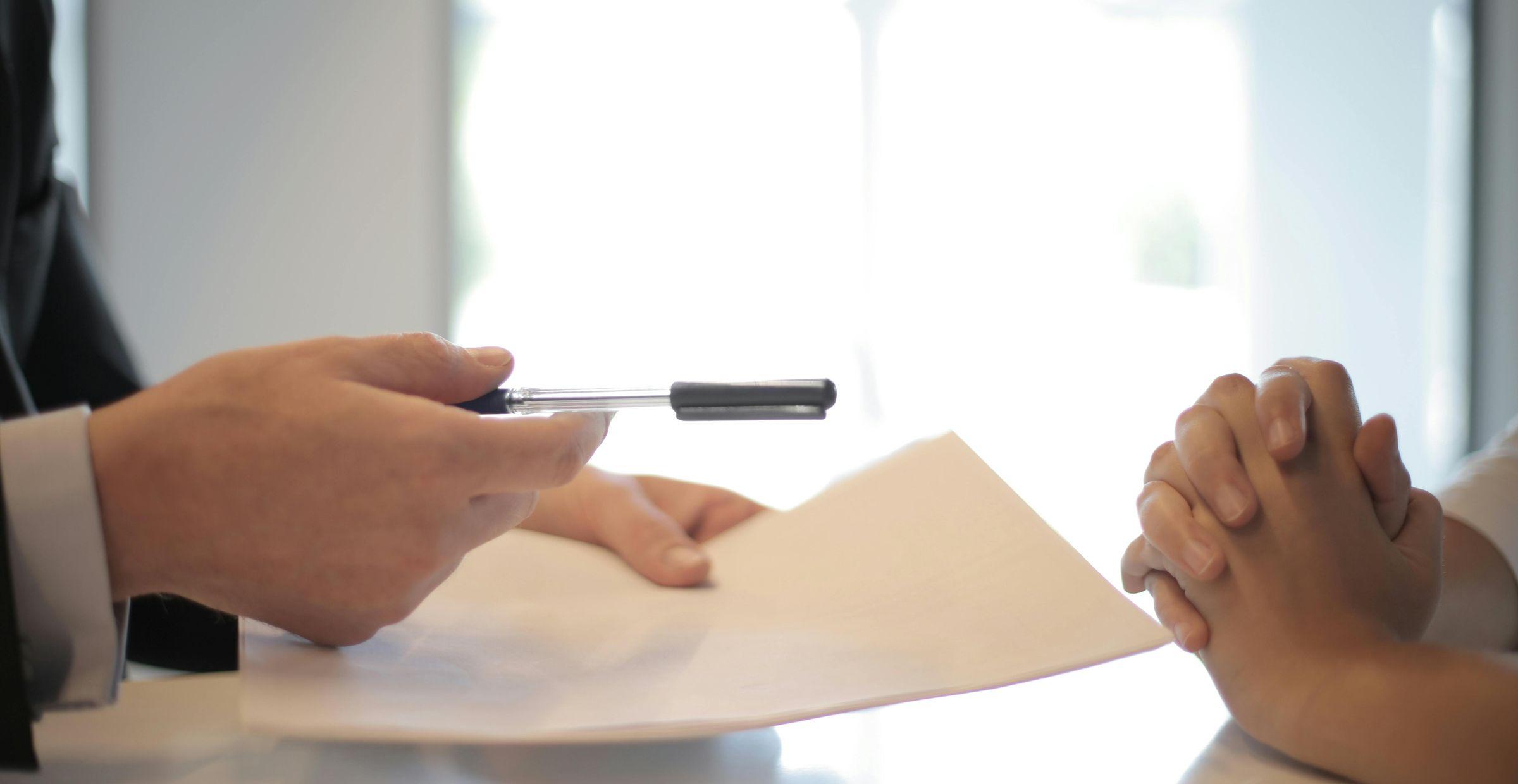 Person überreicht Dokument und Stift zur Unterschrift bei einem Beratungsgespräch.