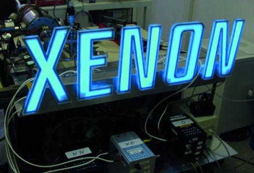 Leuchtendes Schild mit der Aufschrift „XENON“ in blauer Neonbeleuchtung, umgeben von technischen Geräten und Laborinstrumenten.