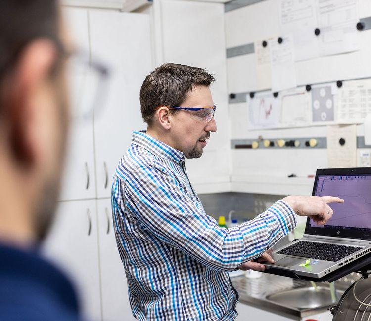 Mann in kariertem Hemd und Schutzbrille arbeitet an einem Laptop in einem modernen Labor mit weißen Schränken und Geräten im Hintergrund